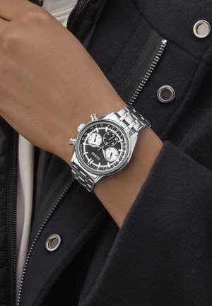 Silver chronograph watch with black dial, white sub-dials, and stainless steel band. Worn on a brown-skinned wrist, contrasting against a dark coat.
