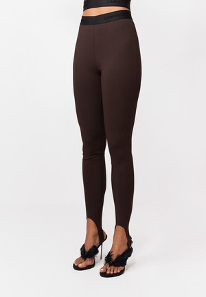 Kobieta modelka w brązowych legginsach z wysokim stanem i detalem na stopę, zestawionych z czarnymi sandałami na wysokim obcasie z piórami.