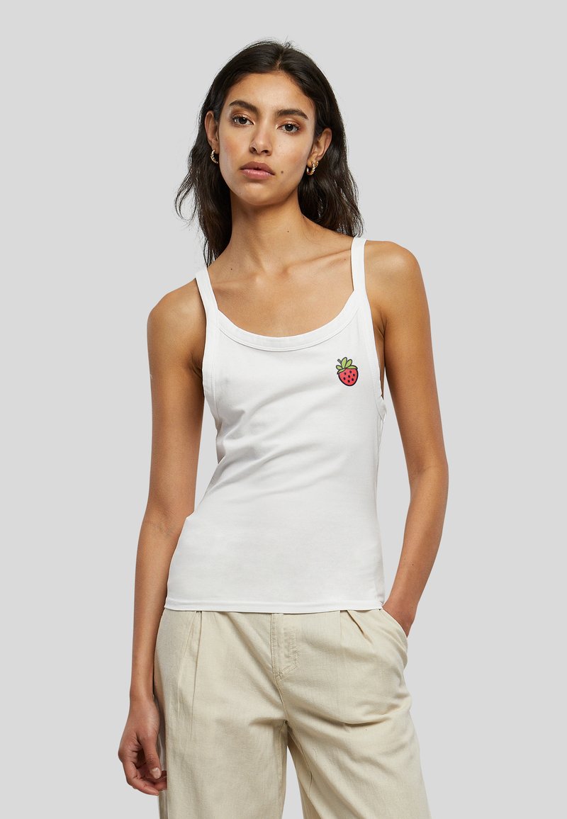 Jeune femme aux longs cheveux foncés porte un débardeur blanc avec un petit motif fraise et un pantalon beige, debout devant un fond uni.