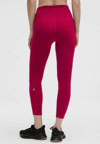 Žena oblečená v svetloružových legínach s vysokým pásom a čiernych športových topánkach, stojaca chrbtom k fotoaparátu na bielom pozadí.