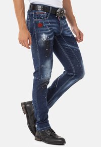 Donkerblauwe denim jeans met versleten details, witte en gekleurde verfspatten, en een rode patch; gecombineerd met zwarte veterschoenen.