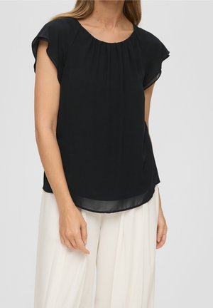 Femme portant un chemisier noir à manches courtes avec un col froncé et un pantalon blanc large sur fond gris.
