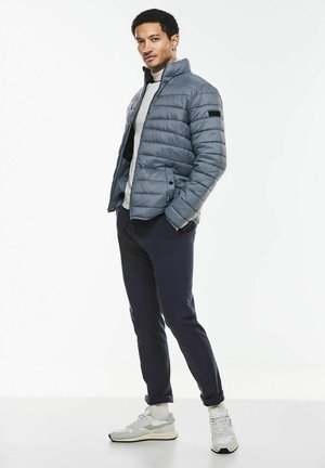 Veste matelassée gris clair avec un col haut, associée à un pull blanc et un pantalon sombre. Portée avec des baskets gris clair et blanches.