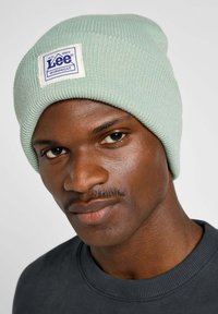 Miętowa dzianinowa czapka z mankietem, z prostokątną białą łatką z niebieskim napisem. Gładka tekstura, dopasowany krój.