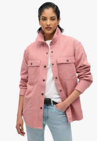 Chemise oversize rose en coton avec col, deux poches poitrine, fermeture à boutons et manches longues. Portée par-dessus un t-shirt blanc.