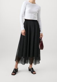 Černá plisovaná midi sukně s volným designem, spárovaná s bílým topem s dlouhými rukávy; černé muly s blokovým podpatkem a malé bordó kabelky.