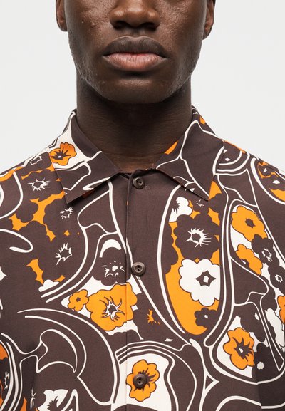Κοντινό πλάνο ενός ατόμου που φορά καφέ πουκάμισο με κουμπιά, με έντονο πορτοκαλί, λευκό και σκούρο λουλουδένιο μοτίβο παϊέτας και μυτερό γιακά.