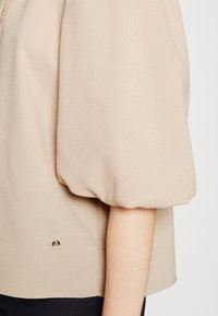 Blus med puffärmar i beige, tillverkad i lätt tyg; har en rundad nedkant och en liten guldbutton detaljerad nära botten.