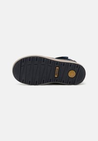 Semelle en caoutchouc noir avec des rainures texturées, semelle intermédiaire beige et un détail de logo circulaire. Conception plate adaptée pour des chaussures décontractées.