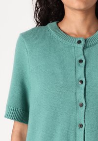 Tyrkysový pletený cardigan s krátkými rukávy, s hebkým výstřihem a šesti černými knoflíky vpředu. Hladká textura a volný střih.