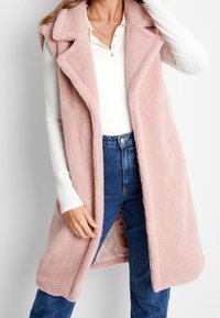 Veste rose sans manches en tissu doux et texturé. Elle présente un col châle et un devant ouvert, associée à une chemise blanche à manches longues et un jean bleu.