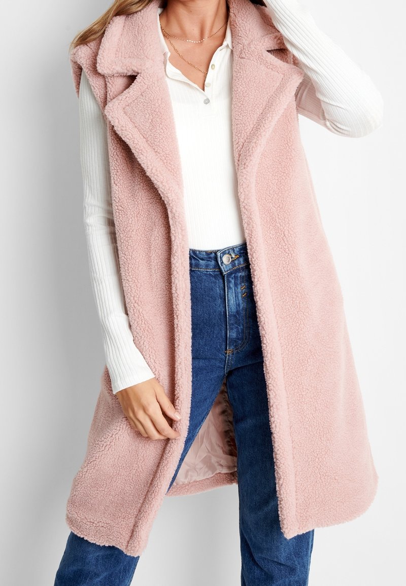 Veste rose sans manches en tissu doux et texturé. Elle présente un col châle et un devant ouvert, associée à une chemise blanche à manches longues et un jean bleu.