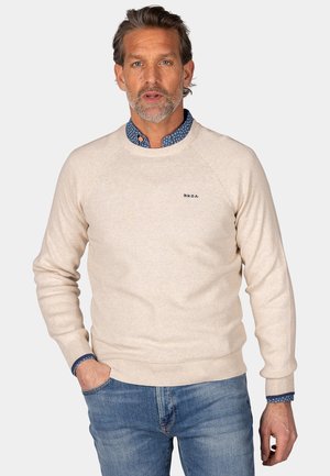 Beige gebreide trui met geribde manchetten en zoom. Heeft een subtiele textuur en een geborduurd logo op de borst. Geddragen over een blauwe overhemd.