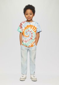 Kolorowa koszulka z wzorem tie-dye w spiralę w odcieniach pomarańczowym, niebieskim i turkusowym. Połączona z jasnoniebieskimi dżinsami i białymi trampkami.