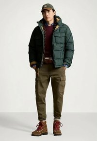 Άνδρας με μακριές πλεξίδες, ντυμένος με πράσινη τζάκετ puffer, μπορντό πουλόβερ, παντελόνια cargo, καφέ μποτάκια πεζοπορίας και πράσινο καπέλο, στέκεται με τα χέρια στις τσέπες.