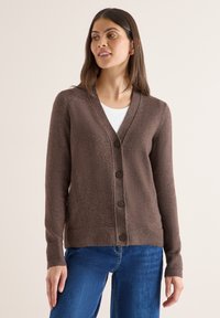 Brun stickad kavaj med V-urringning, fem framknappar och ribbade ärmslut. Bärs över en vit topp och matchas med blå jeans.