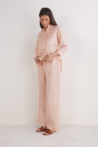 Lichtroze linnenoutfit met een knoopsluiting shirt met een borstzak en losse, met een koord verstelbare broek. Model staat blootsvoets op een zachte ondergrond.