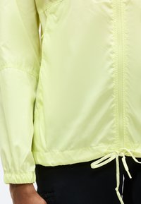 Φωτεινό κίτρινο μπουφάν με φερμουάρ, ελαστικά μανσέτες και ρυθμιζόμενο κορδόνι στη μέση, φορεμένο με μαύρο παντελόνι.