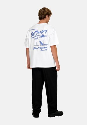 Joven con camiseta blanca con estampado azul de la playa de St. Tropez, pantalones negros y zapatos negros, de pie de espaldas contra un fondo blanco.