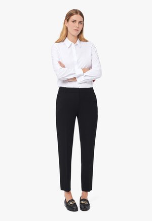Chemise blanche à manches longues, pantalon noir ajusté, et mocassins noirs avec accent en chaîne dorée. Coupe simple et structurée.