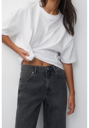 Vrouw tilt lichtjes een oversized wit T-shirt op, draagt een zwarte high-waisted jeans met wassing, tegen een effen witte achtergrond.