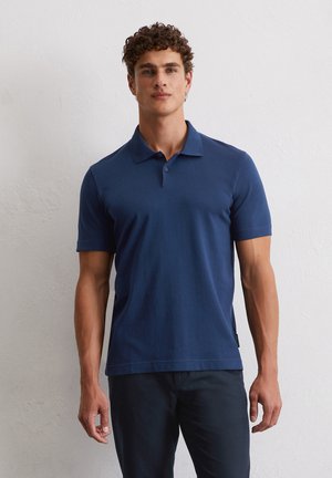 Polo bleu marine en tissu de coton lisse, manches courtes, col boutonné et détails discrets avec fentes latérales. Porté avec un pantalon foncé.