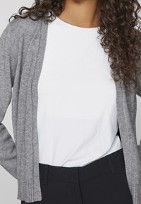 Grå ribbade kavaj med öppen framtill, bärs över en vit skjorta. Har långa ärmar och en mjuk textur. Kombineras med svarta byxor.