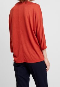 Rustrote, übergroße Pullover mit Dreiviertelärmeln, weicher Textur und einer zentralen Naht auf der Rückseite. Kombiniert mit navyblauen Hosen.
