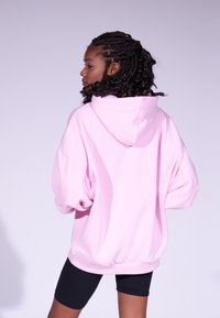 Lys rosa oversized hettegenser med frontlomme og snorhette. Mykt stoff og ribbet mansjetter med avslappet passform. Svart shorts synlig under.