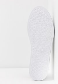 Semelle de chaussure blanche avec un motif diamant texturé, photographiée à la verticale contre un mur blanc et un sol blanc.