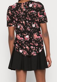 Blusa nera a fiori con maniche corte, caratterizzata da fiori rosa e bianchi, colletto rotondo e dettaglio a chiave sul retro, abbinata a una gonna nera.