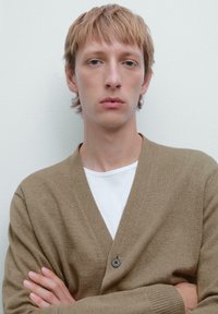 Brun stickad cardigan med V-ringning, enkel knappstängning, bärs över en vit T-shirt, texturerad tyg, armarna i kors mot en neutral bakgrund.