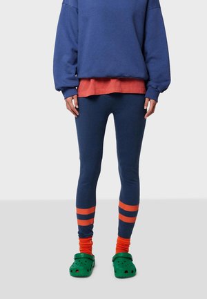 Blauwe oversized sweatshirt gelaagd over een rode top, gecombineerd met marineblauwe legging met oranje strepen en groene klompen. Textuur van de materialen zichtbaar.