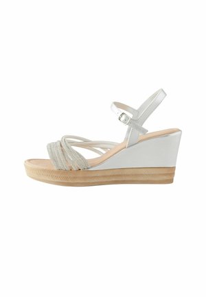 Wedge sandals featuring a white synthetic upper, decorative metallic straps, a light brown woven sole, and an adjustable ankle strap.