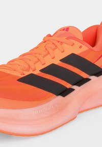 Πορτοκαλί αθλητικό παπούτσι με μαύρο τριών ρίγες σχέδιο, κορδόνια, πλέγμα πάνω μέρος και ροζ μαξιλαρωμένη σόλα με ετικέτα "DREAMSTRIKE GLOW."