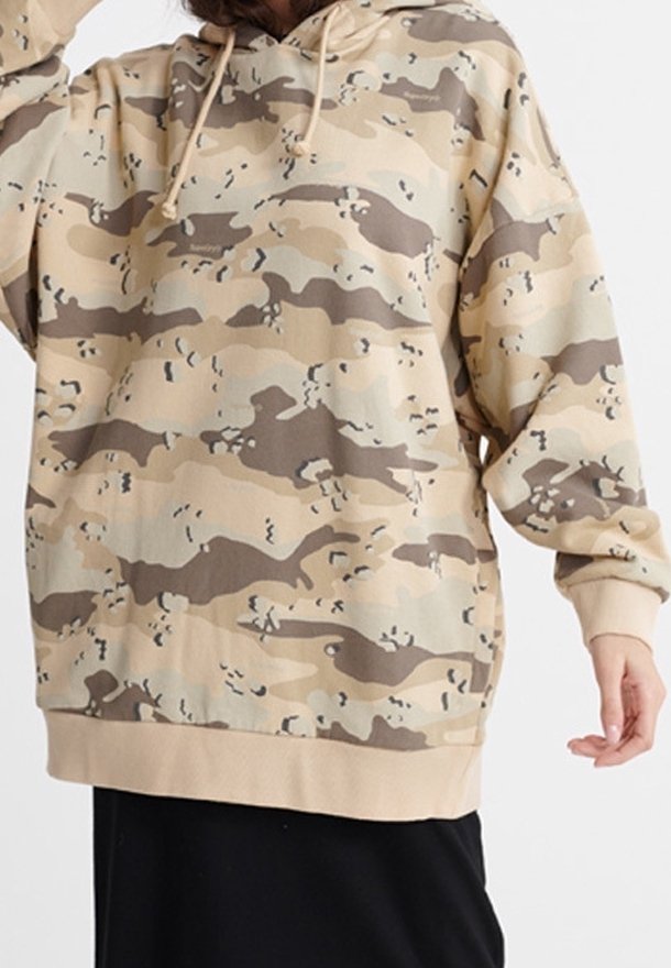 Personne portant un sweat à capuche beige et marron camouflage avec des cordons, associée à une jupe noire, debout devant un fond uni.