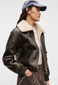 Smeđa kožna jakna s bež umjetnim krznom na ovratniku, zatvaranje na zip, bočne džepove i rebraste manžete; nosi se s plavom kapom i bež majicom.