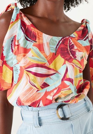 Nainen, joka pukeutuu värikkääseen, olkapäät paljastavaan kukalliseen toppiin, jossa on solmittavat hihnat, yhdistettynä vaaleansinisiin korkeavyötäröisiin farkkuihin ja pyöreällä soljella varustettuun vyöhön.