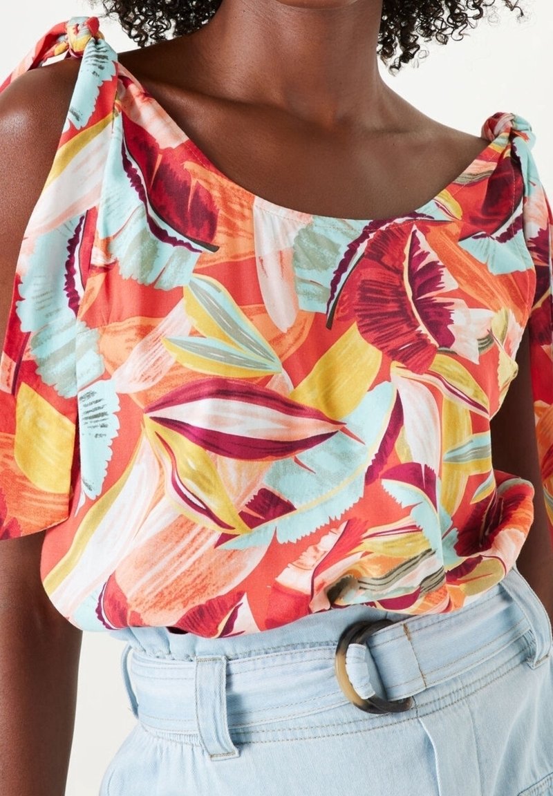 Femme portant un haut coloré à fleurs, épaules dénudées avec des bretelles nouées, associé à un jean taille haute bleu clair et une ceinture à boucle ronde.