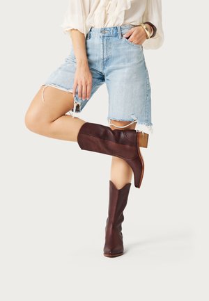 Persoon draagt een crèmekleurige blouse, gescheurde lichtblauwe spijkershorts en bruine leren laarzen tot halverwege de kuit, poseert met een gebogen been.