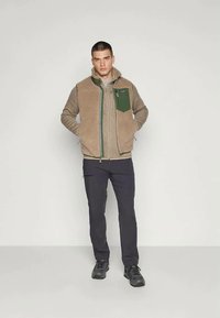 Bărbat în picioare, îmbrăcat cu o jachetă de fleece bej cu buzunar verde pe piept, cămașă gri cu mânecă lungă, pantaloni negri și ghete de drumeție negre.