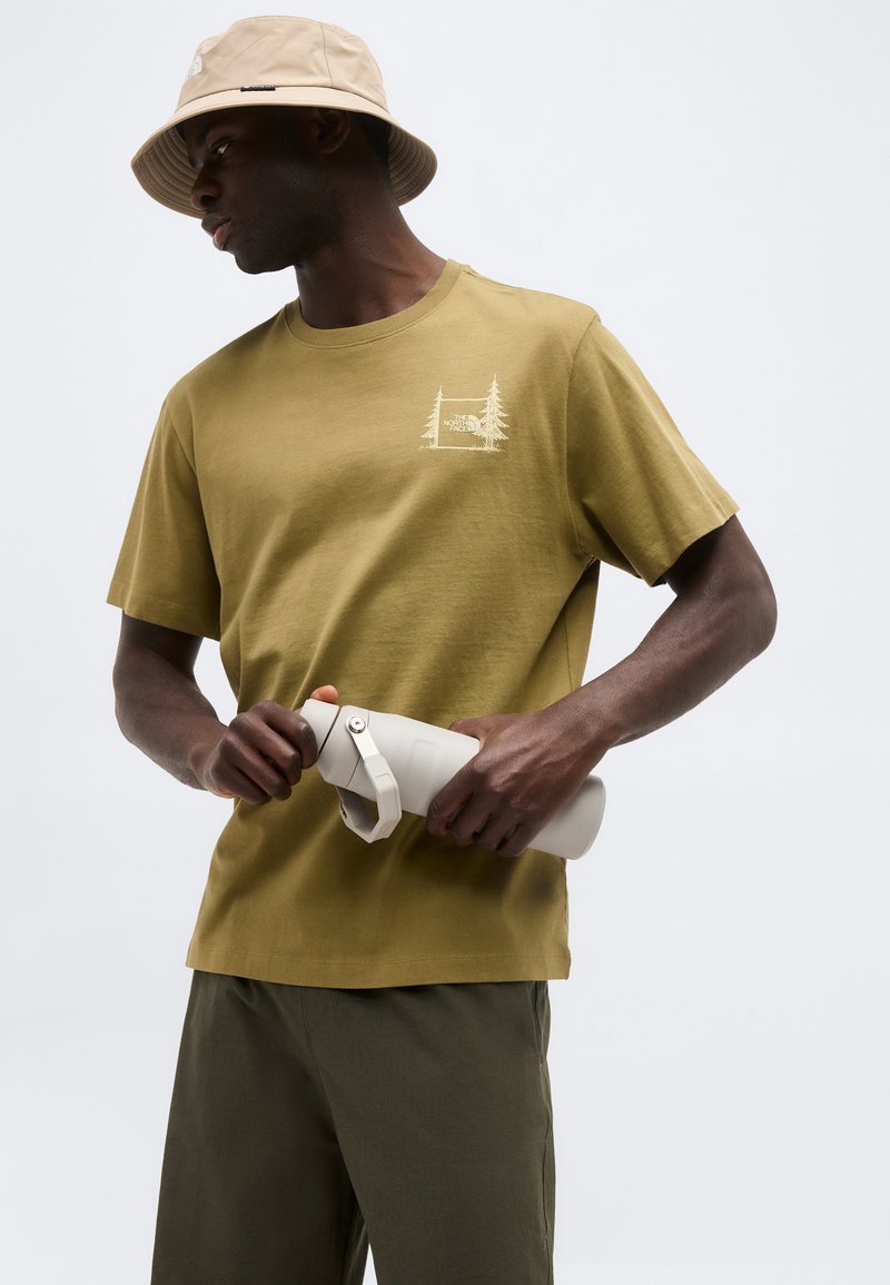 Mann mit beigem Anglerhut und olivgrünem T-Shirt hält eine hellgraue Wasserflasche vor einfarbigem Hintergrund.