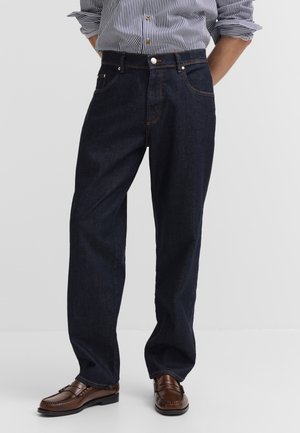 Mann trägt dunkelblaue Jeans mit geradem Bein, ein eingestecktes gestreiftes Hemd mit Knöpfen und braune Lederloafer, steht mit den Händen hinter dem Rücken.