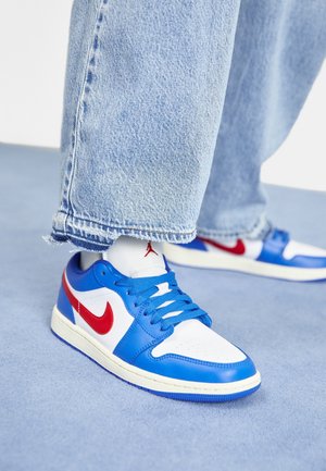 Osoba nosząca niebieskie, białe i czerwone buty Nike z czerwonym logo i niebieskimi sznurowadłami, połączone z jasnoniebieskimi dżinsami na jasnoniebieskiej podłodze.