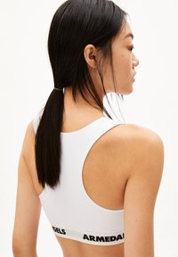 Weißer Sport-BH mit Rundhalsausschnitt und Racerback-Design. Mit einem schwarzen Band mit dem "ARMEDANGELS"-Logo an der Unterseite. Glatte Textur.