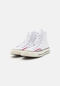 Hvite høye sneakers laget av lerret, med gummisåle, metalløpler og røde og svarte stripeaksenter på siden.