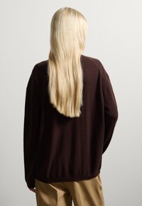 Brauner Strickpullover mit lockerer Passform, langen Ärmeln und abgerundetem Saum, getragen über hellbeigen Hosen. Die Rückansicht zeigt lange, glatte blonde Haare.