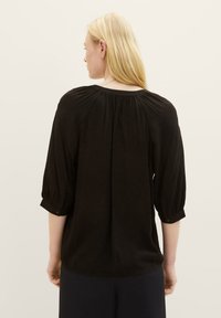 Schwarze Bluse mit drei Viertel Puffärmeln, gerafftem Rundhalsausschnitt und geknöpften Manschetten. Leichter Stoff mit glatter Textur und dezentem Schimmer.