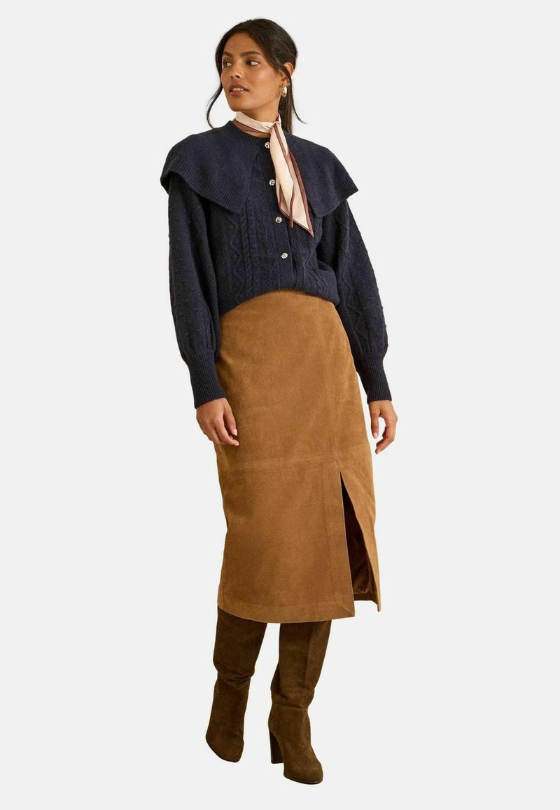 Námornícky pletený sveter s golierom, hnedá zamsková sukňa s predným rozparkom a hnedé čižmy po kolená. Mäkké textúry a vrstvený dizajn.