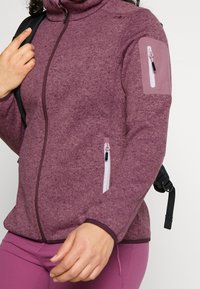 Fleece takki tumman purppurana, jossa on korkea kaulus, olkapatja vetoketjullisella taskulla ja kaksi vetoketjutaskua edessä. Rakenne on neulottua materiaalia.
